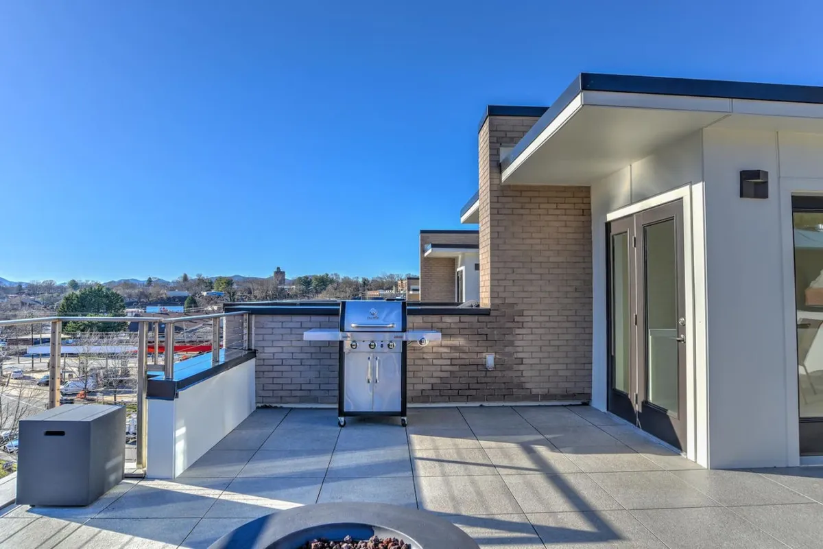 A spacious rooftop terrace featuring a sleek grill station, a modern outdoor seating area with plush cushions, and panoramic views of both the city skyline and distant mountains. The scene is set during golden hour, inviting barbecues and relaxed gatherings.