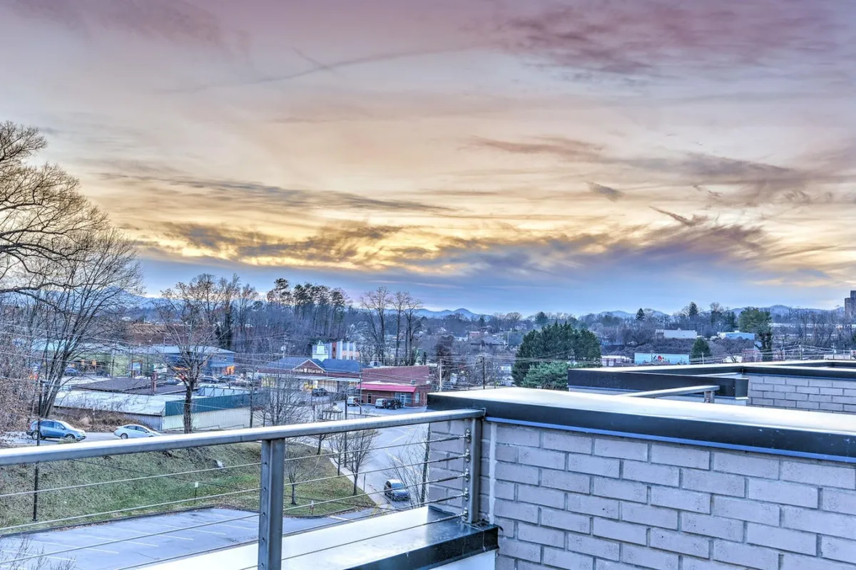 A breathtaking view from the rooftop terrace at sunset, showcasing the glowing sky over the mountain range, urban landscape, and the edge of a balcony railing. The image evokes tranquility and the thrill of being high above city life as evening sets in.