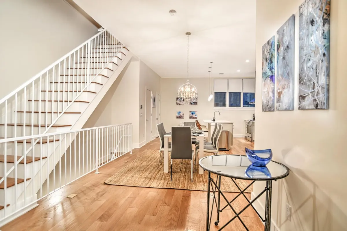 This image showcases a bright and modern dining space with a stylish chandelier, a central dining table, a staircase with clean lines, and carefully selected wall art. The layout suggests a welcoming transition point between the private home and the vibrant downtown environment, inviting both focused work and social interaction.
