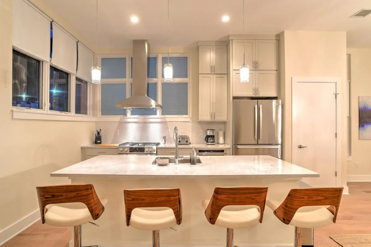 A contemporary kitchen scene with a large island, bar stools, stainless steel appliances, and modern pendant lights. The space is bright and inviting, designed for both socializing and relaxed mornings.