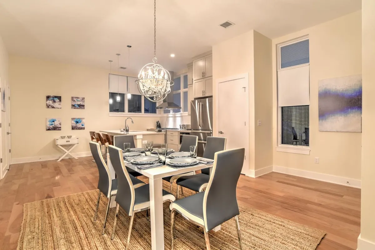 A sophisticated yet welcoming dining area featuring a set table with plates and glasses, a sparkling chandelier, and a kitchen island in the background. The space suggests memorable meals and group gatherings.