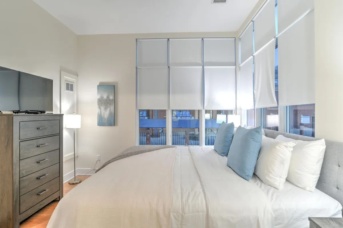 A spacious bedroom featuring a neatly made bed with soft designer linens, large windows letting in natural light, a stylish dresser, an elegant lamp, and framed artwork on the wall. The calming color palette and uncluttered arrangement evoke a sense of tranquility and relaxation.