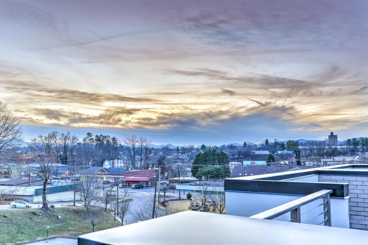 This view-focused image displays a stunning sunset sky over the Asheville cityscape, as seen from a balcony or rooftop perspective. The horizon is bathed in warm light, inviting moments of relaxation and reflection at the close of a day spent exploring downtown.