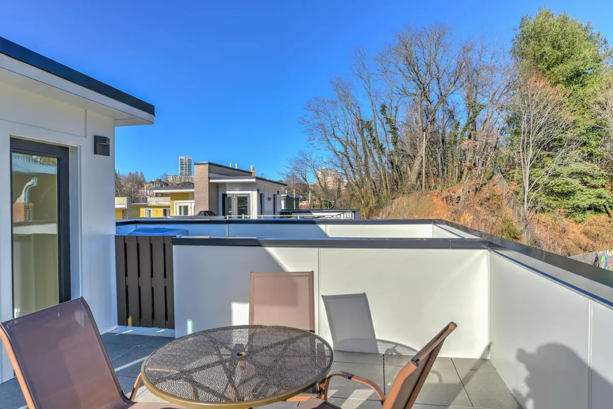 This image captures a stylish outdoor seating area with a glass railing, surrounded by lush trees and featuring a distant view of the cityscape. The space is designed for relaxation, with comfortable chairs oriented to enjoy the natural and urban scenery.
