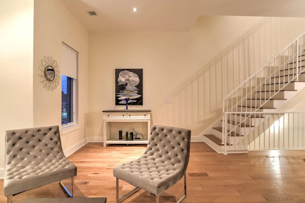 A sophisticated interior vignette with two tufted chairs set for conversation, abstract artwork on the walls, and a modern staircase rising behind them. The mood is serene and refined, ideal for unwinding or greeting guests.