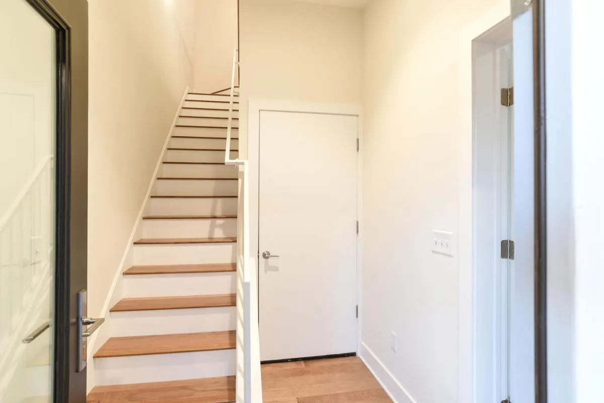 A bright, inviting entryway with a staircase ascending from a white door, surrounded by light-colored walls. The scene evokes a sense of welcome and the beginning of a comfortable stay.